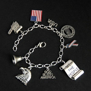 Op de afbeelding: Zilveren armband met charmes met een patriottisch thema, waaronder een bel, een adelaar, een rol met de tekst "We the People", een vlag en een lint. De armband heeft ook charmes met de tekst "God Bless America" en "Home of the Brave".