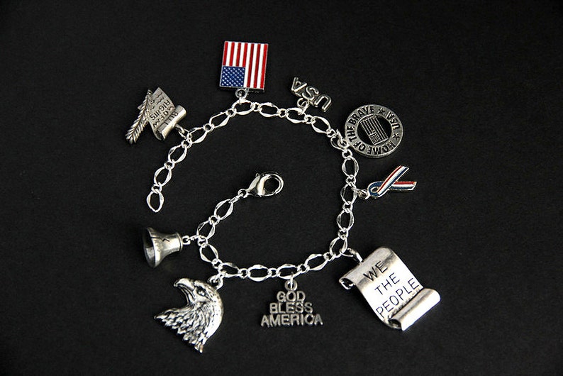 Op de afbeelding: Zilveren armband met charmes met een patriottisch thema, waaronder een bel, een adelaar, een rol met de tekst "We the People", een vlag en een lint. De armband heeft ook charmes met de tekst "God Bless America" en "Home of the Brave".