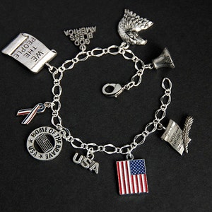 Op de afbeelding: Een zilveren armband met verschillende patriottische bedels, waaronder een rol met de tekst "We the People", een lint, een bel, een Amerikaanse zeearend, een cirkel met de tekst "Home of the Brave" en een ster, een bedel met de tekst "USA" en een bedel met een Amerikaanse vlag.