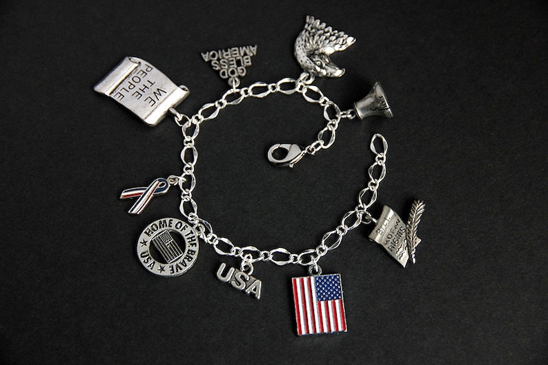 Op de afbeelding: Een zilveren armband met verschillende patriottische bedels, waaronder een rol met de tekst "We the People", een lint, een bel, een Amerikaanse zeearend, een cirkel met de tekst "Home of the Brave" en een ster, een bedel met de tekst "USA" en een bedel met een Amerikaanse vlag.