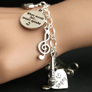 May include: Silver charm bracelet with music-themed charms, including a treble clef, a microphone, and a heart charm that says "Singing". The round charm says "When words fail... music speaks".