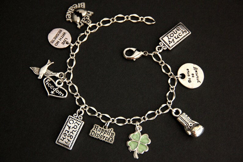 May include: A silver charm bracelet with multiple charms, including a heart with the word "Freedom" on it, a bird, a four-leaf clover, a boxing glove, and several rectangular charms with text. The text on the charms includes "Live with no regrets", "Hear Breaker", "Laugh Often", "Love Much", "Happily Divorced", and "Be true to yourself".
