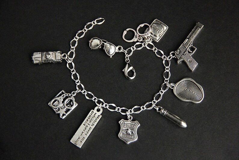 May include: Silver charm bracelet with various charms including a police badge, handcuffs, a gun, sunglasses, a fingerprint, a bullet, and a car. The bracelet also has a charm with the text "Watch over me while I protect and serve."