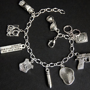 May include: Silver charm bracelet with various police-themed charms, including handcuffs, a badge, a gun, a fingerprint, sunglasses, and a bullet. The bracelet also has a charm with the text "Watch over me while I serve and protect."