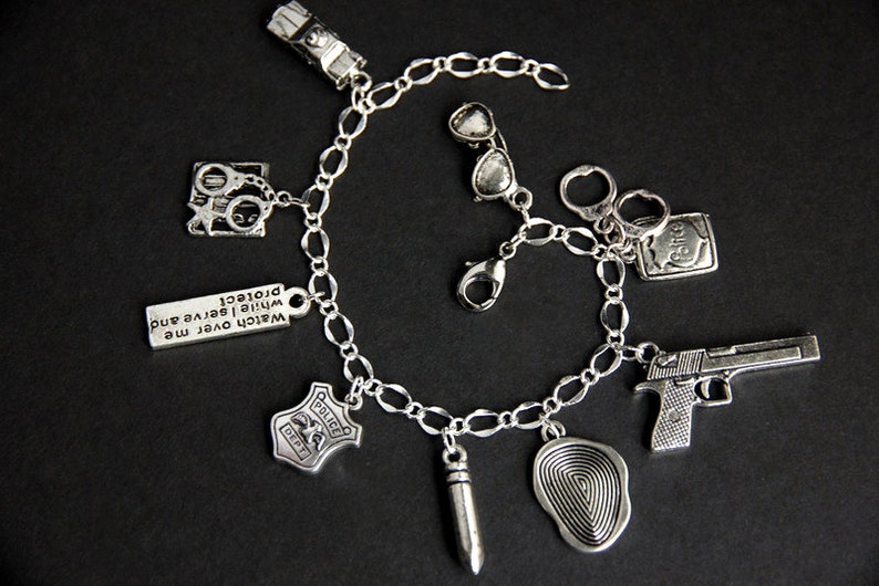 May include: Silver charm bracelet with various police-themed charms, including handcuffs, a badge, a gun, a fingerprint, sunglasses, and a bullet. The bracelet also has a charm with the text "Watch over me while I serve and protect."