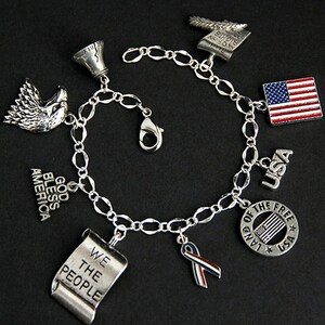 Op de afbeelding: Een zilveren armband met bedels met een patriottisch thema, waaronder een adelaar, een bel, een rol met de tekst "We the People", een lint, een vlag en een cirkel met de tekst "Land of the Free" en een kleine vlag erin.