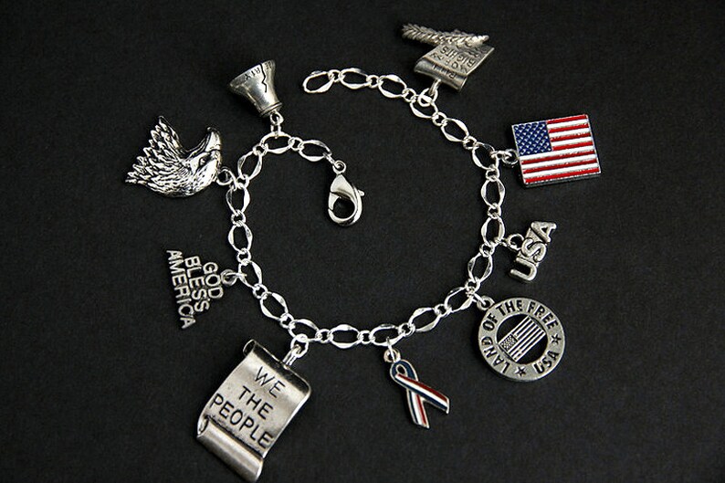 Op de afbeelding: Een zilveren armband met bedels met een patriottisch thema, waaronder een adelaar, een bel, een rol met de tekst "We the People", een lint, een vlag en een cirkel met de tekst "Land of the Free" en een kleine vlag erin.