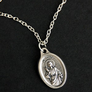 May include: A silver chain necklace with a silver oval pendant featuring a depiction of Saint Lucy. The pendant has the text "St. Lucy" inscribed on it.