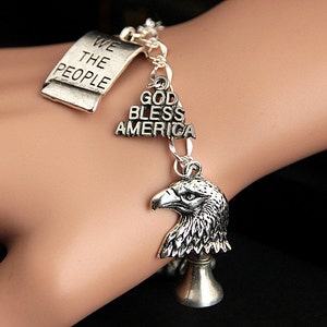 Op de afbeelding: Een zilveren armband met meerdere bedels, waaronder een tag met de tekst "We the People", een bedel met de tekst "God Bless America" en een bedel van een adelaarskop.