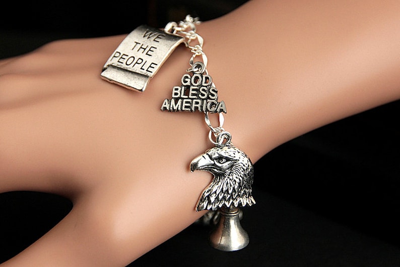 Op de afbeelding: Een zilveren armband met meerdere bedels, waaronder een tag met de tekst "We the People", een bedel met de tekst "God Bless America" en een bedel van een adelaarskop.