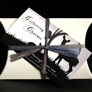 May include: A white pillow box with a black and white business card tied with a blue ribbon. The business card says "Gathering Charms" and has a silhouette of a deer in a forest. The business card also includes the address "4928, S. 1st Ave, Tacoma, WA 98295" and the email address "gatheringcharms@gmail.com".