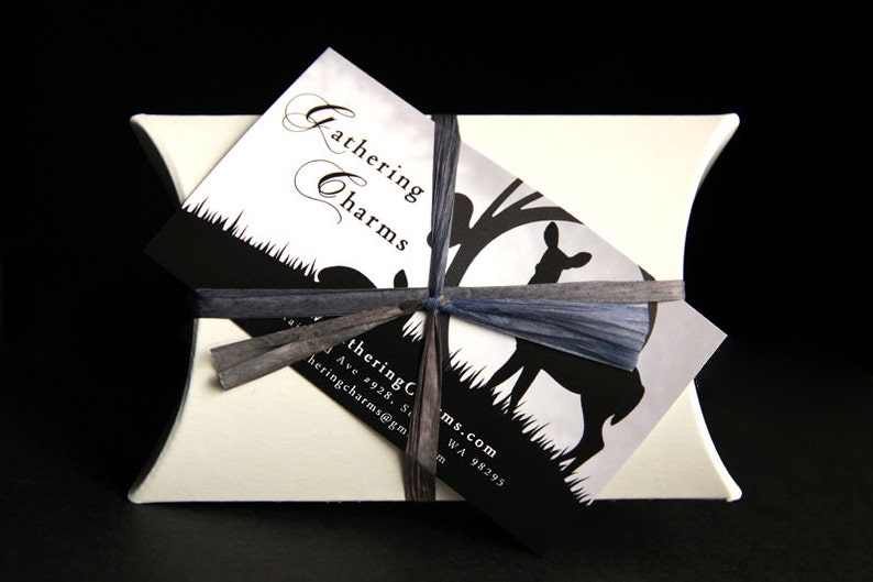 May include: A white pillow box with a black and white business card tied with a blue ribbon. The business card says "Gathering Charms" and has a silhouette of a deer in a forest. The business card also includes the address "4928, S. 1st Ave, Tacoma, WA 98295" and the email address "gatheringcharms@gmail.com".