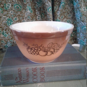 Puede incluir: Un tazón de mezcla marrón y blanco con un diseño de fruta. El tazón está sobre un libro azul que dice "The American Woman's Cook Book".