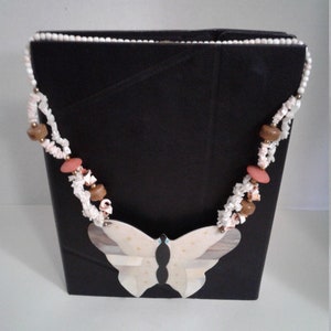 Peut inclure: Un collier avec un pendentif en forme de papillon blanc avec des perles brunes et roses. Le collier est sur un fond noir.