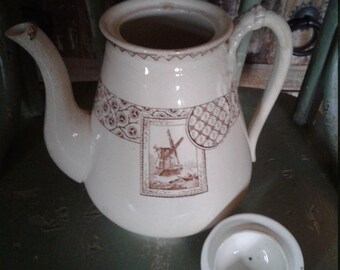 May include: A white ceramic teapot with a brown windmill design and a matching lid. The teapot has a round body and a spout on the side. The lid has a small knob on top.