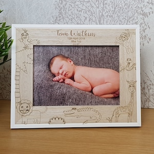 May include: A white wooden photo frame with a light brown woodgrain finish. The frame has a cutout design of jungle animals, including a giraffe, lion, monkey, and crocodile. The frame has a white mount and a clear glass front. The frame is displaying a photo of a baby sleeping on a grey blanket. The photo frame has the text "Tom Watkins 12th April 2019 8lbs 10oz 19:47" engraved on the front.