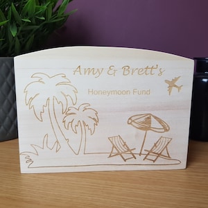 May include: A light-colored wooden box with the engraved text "Amy & Brett's Honeymoon Fund." The box features etched images of palm trees, a beach scene with lounge chairs, an umbrella, and an airplane.