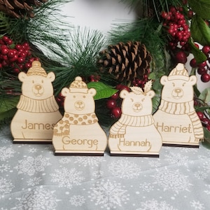Könnte beinhalten: Vier Holz-Platzkartehalter in Form von Eisbären mit den Namen "James", "George", "Hannah" und "Harriet" auf einer weißen Tischdecke mit Schneeflockenmuster.