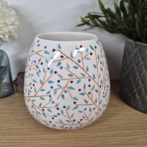 Könnte beinhalten: Eine weiße Keramikvase mit runder Form. Die Vase ist mit einem Muster aus braunen Ästen, hellblauen Blättern und kleinen blauen und roten Punkten verziert. Die Vase steht auf einer Holzoberfläche.