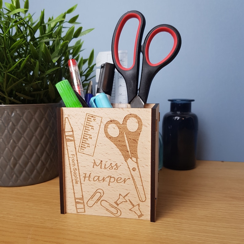 May include: A wooden pencil holder with a personalized inscription that reads "Miss Harper" and a design of school supplies including a ruler, scissors, crayons, paper clips, and push pins. The holder is filled with pens, pencils, and scissors.