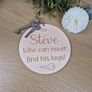 May include: A circular wooden keyring engraved with the words "For Steve Who can never find his keys!" and a key outline. Two keys are attached. The keyring is light brown with dark brown text.