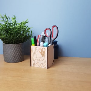 May include: A wooden pencil holder with a personalized inscription "Miss Harper" and a design of school supplies, including a ruler, scissors, paper clips, and a pencil. The holder is filled with pens, pencils, and markers.