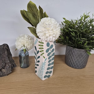 May include: A patterned vase with white flowers and green leaves. A smaller vase with a white flower is to the left. A potted plant in a grey pot is to the right. The vase is on a wooden surface.