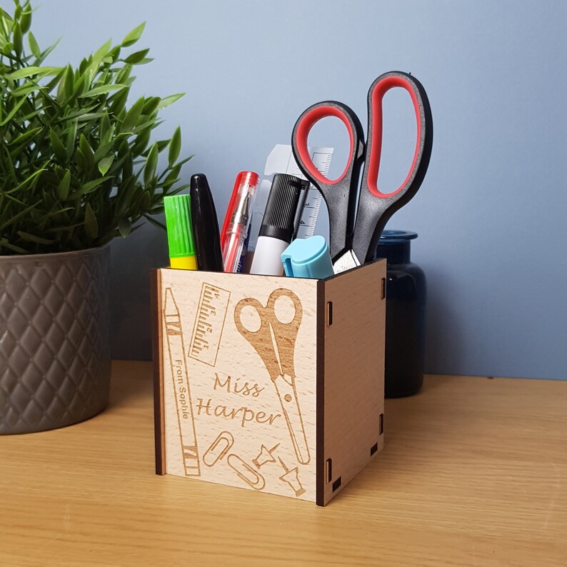 May include: A wooden pencil holder with a personalized message that reads "Miss Harper" and includes a ruler, scissors, paper clips, and a crayon. The holder is engraved with the message "From Sophie".