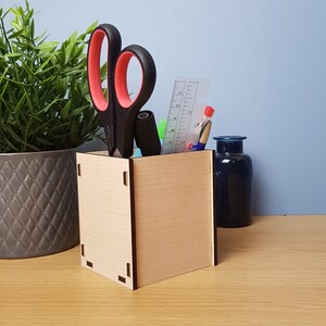 May include: A wooden desk organizer with a natural wood finish. It is filled with office supplies, including a pair of black scissors with pink handles, a ruler, a blue pen, and a black pencil.