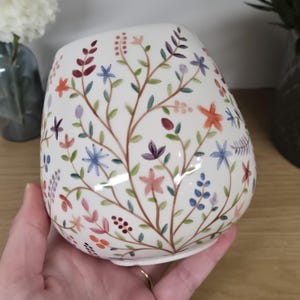 Peut inclure: Vase en céramique blanche orné d'un motif floral. Le vase présente un motif peint à la main de fleurs et de feuilles colorées sur des tiges brunes. Les fleurs sont dans des tons de rouge, orange, rose, bleu et violet. Le vase a une forme arrondie.