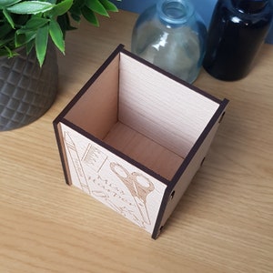 May include: A wooden desk organizer with a dark brown finish. The organizer has a square shape and is decorated with a laser-engraved design of scissors, a pencil, and the text "Must Have Supplies".