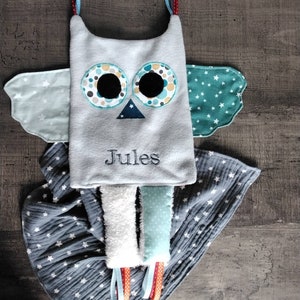 May include: A soft, gray owl-shaped baby toy with embroidered details and the name "Jules". The owl has large, round eyes with colorful accents, wings, and dangling ribbons. It is paired with a gray blanket with white star patterns.