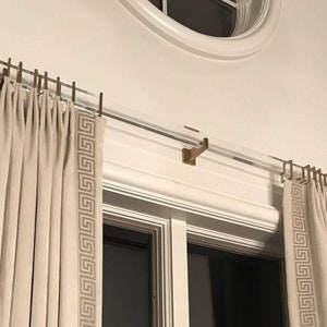 May include: A pair of white curtains with a geometric pattern border hang from a gold metal curtain rod. The rod is mounted to the wall above a window.