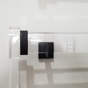 May include: A clear acrylic shelf with black metal brackets. The shelf is mounted to a white wall.