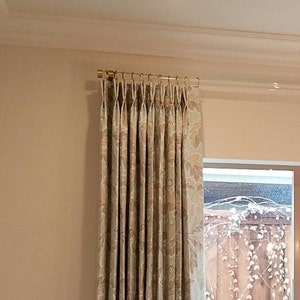 May include: Beige curtains with a floral pattern hang from a gold curtain rod. The curtains are pleated and have a subtle sheen. The curtains are in front of a window with a view of a backyard.