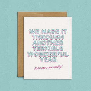 May include: A white card with pink and blue text that reads "We made it through another terrible wonderful year. Let's pop some bubbly!"