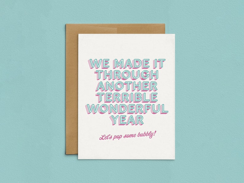 May include: A white card with pink and blue text that reads "We made it through another terrible wonderful year. Let's pop some bubbly!"