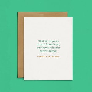 May include: A white greeting card with a brown envelope. The card features green text that reads, "That kid of yours doesn't know it yet, but they just hit the parent jackpot." Below this, yellow text reads, "CONGRATS ON THE BABY!"
