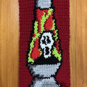 Puede incluir: Un rectángulo rojo tejido a mano con un diseño de lámpara de lava. La lámpara de lava es gris, negra, roja, verde y blanca, con un gráfico de calavera. El fondo es rojo liso y texturizado.