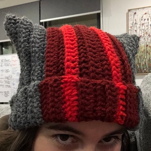 May include: A grey and red crocheted hat with cat ears. The hat has red stripes on the top and grey on the sides.