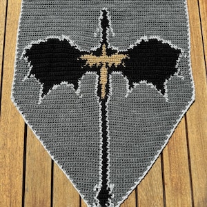 May include: A gray crocheted tapestry with a black and gold dragon design. The tapestry is hanging on a wooden wall.