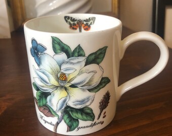 May include: White ceramic mug with a floral design featuring a large white magnolia flower with green leaves and a butterfly. The text "Magnolia grandiflora" is printed on the bottom of the mug.