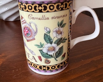 May include: A white ceramic mug with a yellow background and a floral design. The mug has a black and gold border and the text "Camellia sinensis" is printed on the front. The mug is decorated with a teapot, flowers, and leaves.