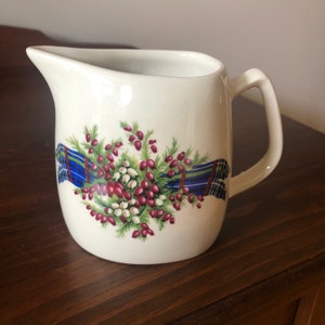May include: White ceramic milk jug with a floral design featuring red berries, green leaves, and a blue and white tartan ribbon.
