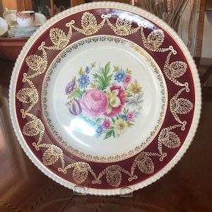 May include: A white porcelain dinner plate with a burgundy rim and gold trim. The center features a floral design with pink, yellow, and blue flowers.