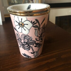 Op de afbeelding: Een roze keramische mok met een gouden rand en een zwart-wit bloemenontwerp.