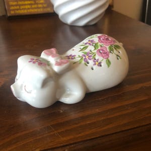May include: A white ceramic pig figurine with pink flowers and a pink bow. The pig is lying on its side with its eyes closed.