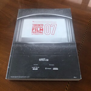 May include: A black and white image of a movie theater with a large screen displaying the text "Toronto International Film Festival 07" and the dates "September 6 to 15". The screen is surrounded by red curtains. The theater is empty, with rows of seats visible in the foreground. The text "tiff07.ca" is displayed at the bottom of the image.