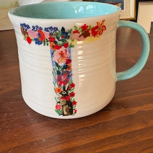 May include: White ceramic mug with a light blue interior and handle. The mug features a floral design in the shape of the letter "T" with a rabbit illustration. The mug has a ribbed texture.