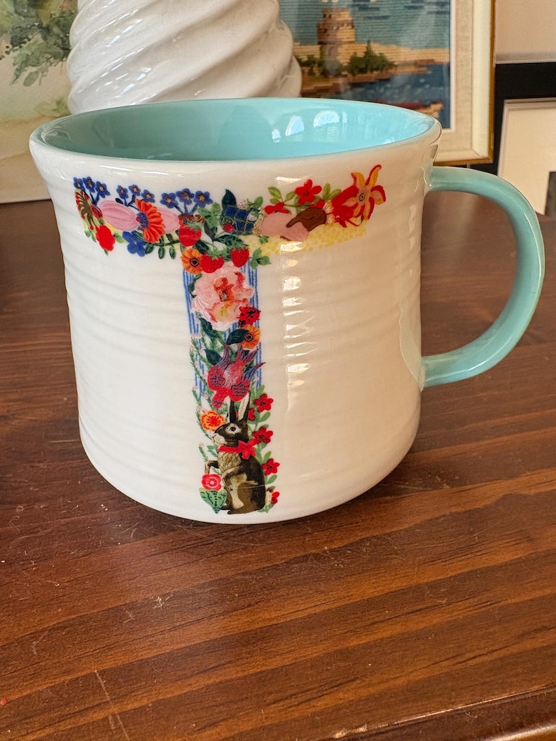 May include: White ceramic mug with a light blue interior and handle. The mug features a floral design in the shape of the letter "T" with a rabbit illustration. The mug has a ribbed texture.